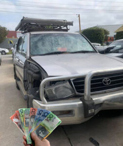 Silver Toyota 4WD with front damage and roof rack; fanned Australian dollar notes shown in hand