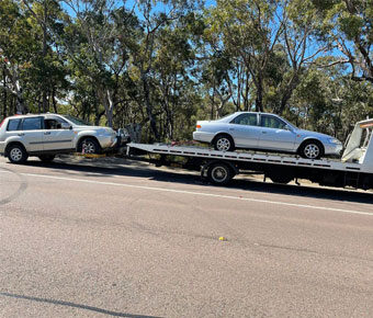 A flatbed tow truck performing a car removal service, loading a silver sedan on a roadside with trees in the background.