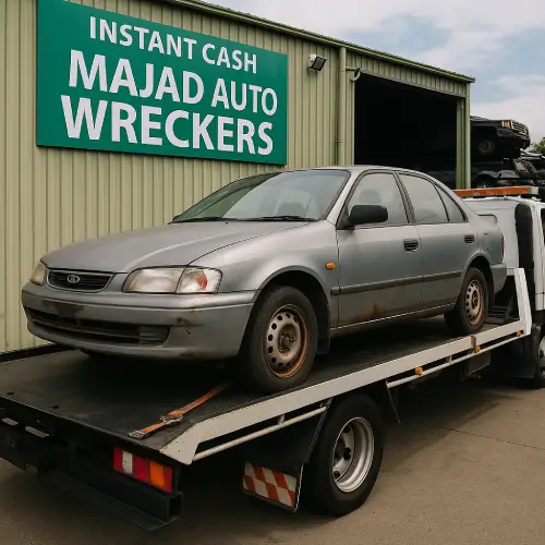 A worn silver sedan being loaded onto a tow truck outside an auto wrecking facility